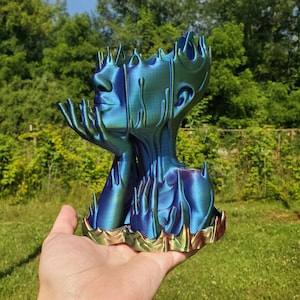 Puede incluir: Un macetero de color azul y verde iridiscente impreso en 3D con forma de cabeza humana con las manos levantadas a los lados. El macetero tiene un borde dorado.