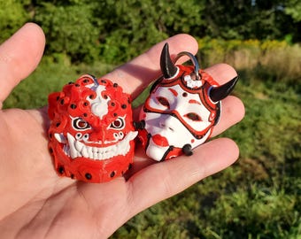 May include: Two red and white 3D printed masks with black details. One mask is a stylized demon face with fangs and the other is a stylized female face with horns. Both masks have a silver ring attached.