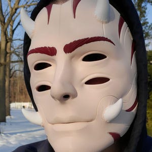May include: A cream-colored mask with white horns and red accents. The mask has eye and mouth openings, and is worn with a black hooded sweatshirt. The mask has a smooth finish and is designed to resemble a face.