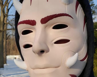 May include: A cream-colored mask with white horns and red accents. The mask has eye and mouth openings, and is worn with a black hooded sweatshirt. The mask has a smooth finish and is designed to resemble a face.