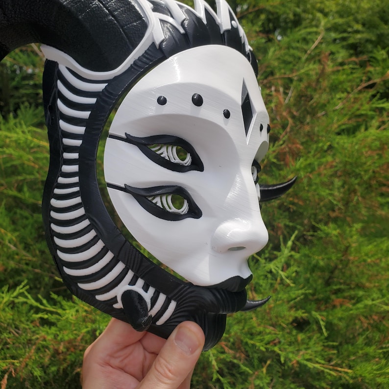 May include: A white and black 3D printed mask with a horned headdress. The mask has a stylized human face with three eyes and a black mouth.