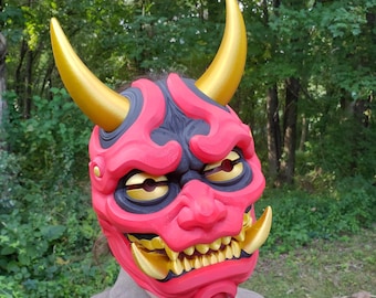 May include: A red and black Oni mask featuring two large, smooth gold horns extending from the top and two smaller gold fangs protruding from the lower jaw. The mask has deep-set gold eyes, a prominent nose, and a wide, toothy grin with gold teeth.