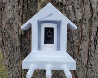 May include: White birdhouse with a Ring security camera mounted inside. The birdhouse has a small bird silhouette on the roof and a feeding tray below the camera.