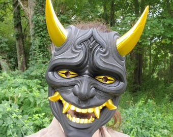 May include: A black Hannya mask featuring prominent, smooth, metallic gold horns extending upward from the forehead. The mask displays intricate, carved-style facial details, including sharp gold teeth and narrow, almond-shaped gold eyes.