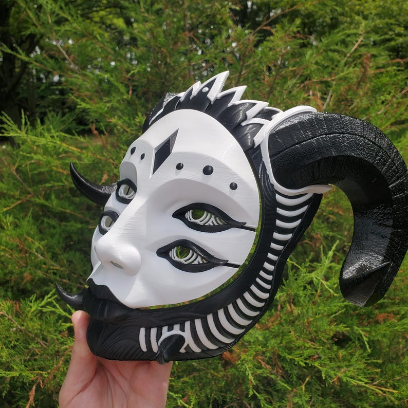 May include: A white and black 3D printed mask with horns, three eyes, and intricate details. The mask has a stylized human face with a black diamond on the forehead.