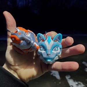 May include: Two cat-shaped keychains. One is gray and orange, the other is blue, white, and gray. The keychains have a layered design and are held in a hand. The cat faces have detailed features.
