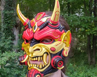 May include: A red, gold, and black Oni mask featuring prominent curved horns, sharp fangs, and mechanical-style circular details on the cheeks. The mask covers the upper face and nose, showcasing intricate sculpted ridges and a vibrant color scheme.