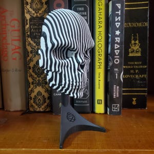 May include: A black and white striped 3D printed skull sculpture on a black stand. The skull is made of many layers of stripes that create a 3D effect.