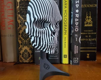 May include: A black and white striped 3D printed skull sculpture on a black stand. The skull is made of many layers of stripes that create a 3D effect.