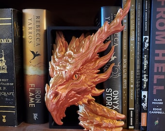 May include: A metallic orange dragon head bookend with intricate details, including an open mouth and a focused eye. The bookend is placed in front of a collection of books, adding a fantasy element to a bookshelf.