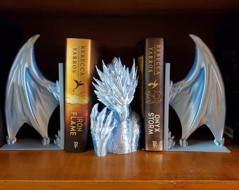 May include: A set of fantasy-themed bookends featuring a dragon design. The bookends include two light blue dragon wings and a silver dragon head. The bookends are holding two books with gold and brown covers.