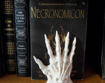 May include: White skeletal hand bookend with sharp claws. The bookend is in front of a black book with gold lettering that reads "Commemorative Edition NECRONOMICON".