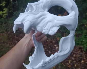 May include: White plastic skull mask with an open jaw and visible teeth. The mask is held in a hand with the jaw open.