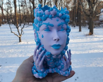 May include: A blue and purple ceramic planter shaped like a woman's head with bubble-like details. The planter has a face with closed eyes and a slightly open mouth.