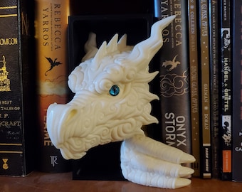May include: A cream-colored dragon head sculpture with intricate details, including horns, scales, and a single blue eye. The dragon is positioned in front of a shelf of books, creating a fantasy theme.