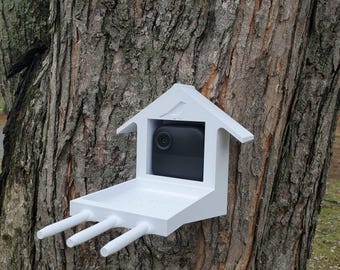 May include: A white bird feeder with a built-in camera, mounted on a tree trunk. The feeder has a flat platform and three perches.