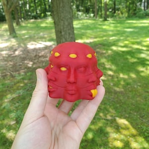 May include: A red sculpture with multiple faces and yellow eyes. The sculpture is held in a hand against a backdrop of green grass and trees. The faces are detailed with lips and noses.
