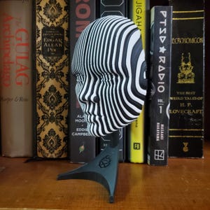 May include: A black and white striped 3D printed sculpture of a face on a black stand. The sculpture is made of multiple layers of stripes that create a 3D effect.