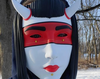 May include: A white mask with red accents, black hair, and white horns. The mask features a red eye covering, black eyebrows, and red lips. Two small gold dots are on the mask. The mask is worn over a blue hooded sweatshirt.