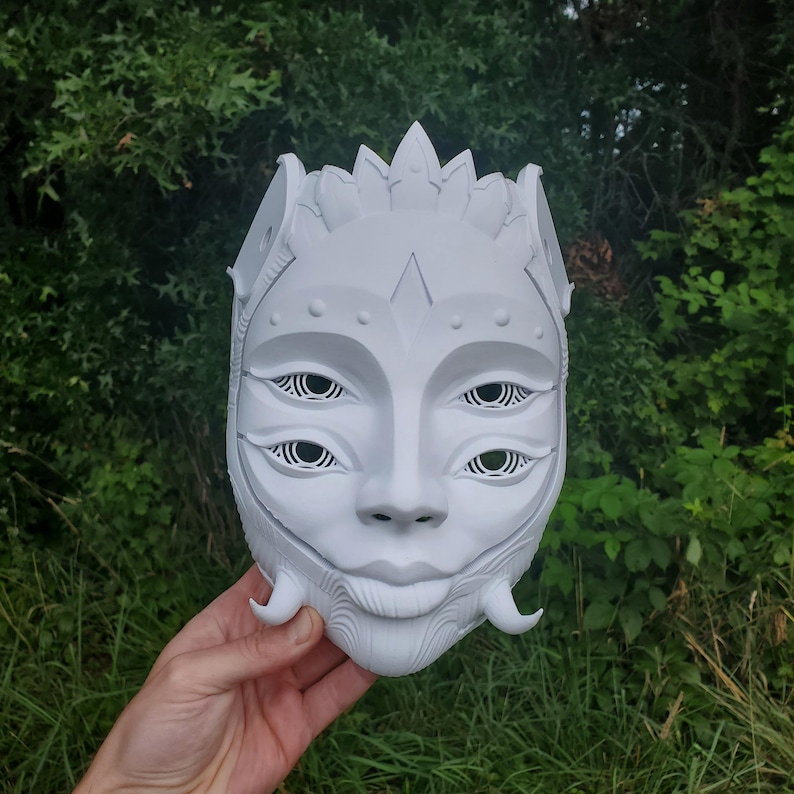 May include: White mask with a detailed design, featuring a face with three sets of eyes and a crown-like headpiece.