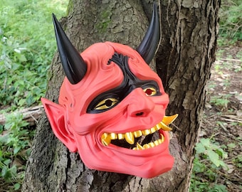 May include: A red and black Oni mask with gold teeth. The mask has a fierce expression and is made of a hard plastic material.