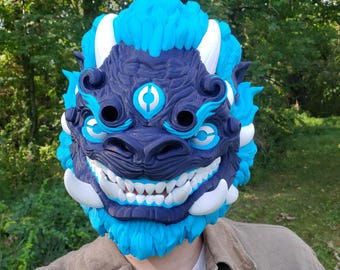 May include: A full-face mask featuring a stylized creature design with a dark navy blue central face, bright cyan blue mane and accents, and white horns and teeth. The mask includes a third eye symbol on the forehead and large eye openings.
