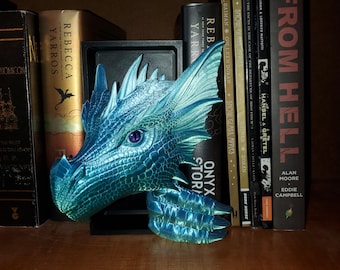 May include: A teal and blue dragon head sculpture with a purple eye, resting on a wooden shelf. The dragon is detailed with scales and fins. Books are visible in the background, including titles like "From Hell" and "Rebecca Yarros."