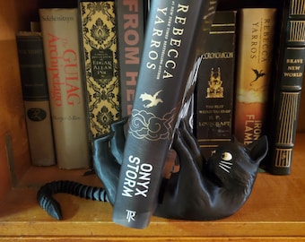 May include: A black cat-shaped bookend holds a book titled "Onyx Storm" by Rebecca Yarros. The bookend is on a wooden shelf with other books.