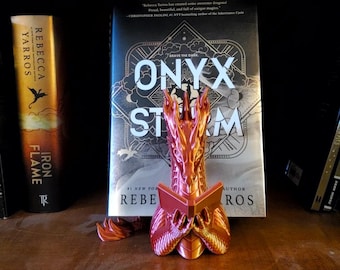 May include: A metallic orange and pink dragon bookend is displayed in front of a book titled "ONYX STORM". The bookend is a detailed sculpture of a dragon holding a book. The bookend is on a wooden shelf with other books.