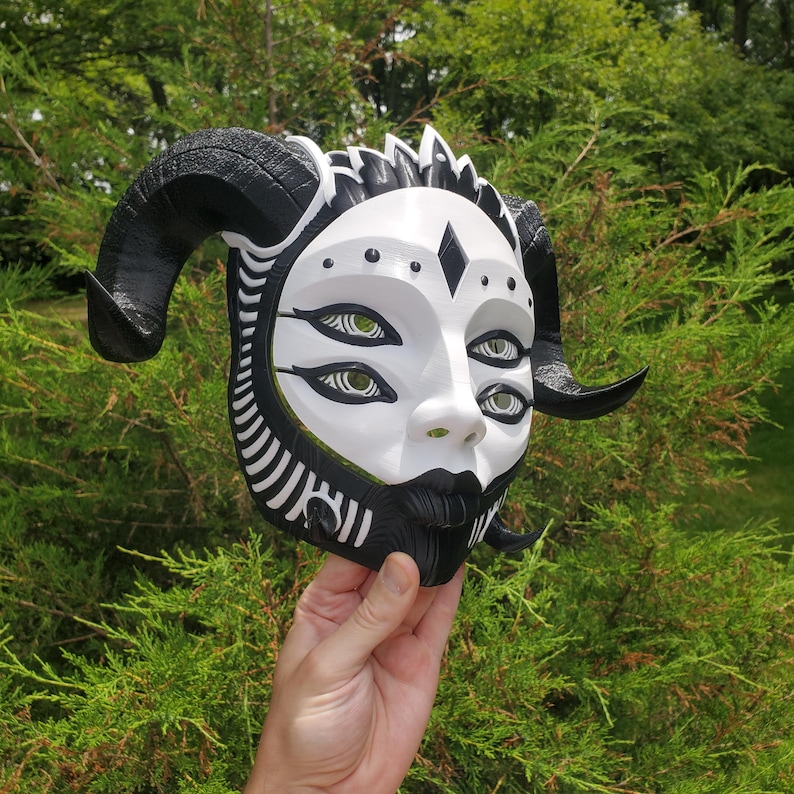 May include: A black and white mask with horns and four eyes. The mask is made of a smooth, white material and has a black and white pattern around the edges. The eyes are painted black with white pupils.