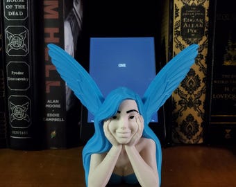 Puede incluir: Una figurita de hada azul y blanca con alas, sentada con la barbilla apoyada en las manos. El hada tiene el pelo azul y mira a la cámara. La figurita está sentada en un estante frente a una estantería. La palabra "ONE" es visible en una pantalla detrás de la figurita.
