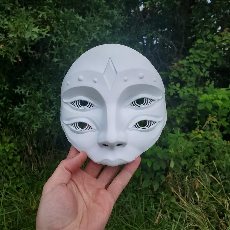May include: White ceramic mask with a human-like face and four eyes. The mask has a diamond-shaped design on the forehead and a series of small circles around the eyes.