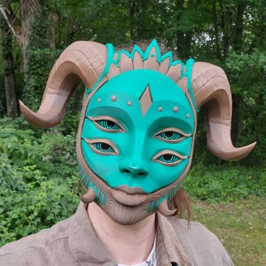 May include: A teal-colored mask featuring four eyes with concentric circular patterns, a central diamond-shaped forehead detail, and two large, curved brown horns extending from the sides. The mask includes intricate textured brown accents.