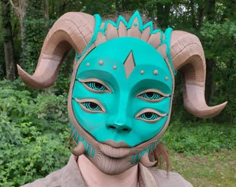May include: A teal-colored mask featuring four eyes with concentric circular patterns, a central diamond-shaped forehead detail, and two large, curved brown horns extending from the sides. The mask includes intricate textured brown accents.