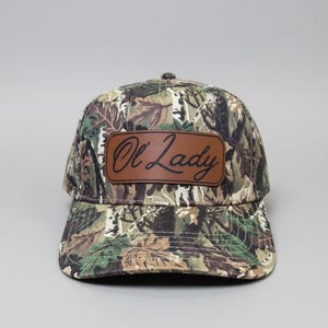 May include: A camouflage baseball cap with a brown leather patch. The patch features the words "Ol' Lady" in a cursive font. The cap has a curved brim and a button on top.