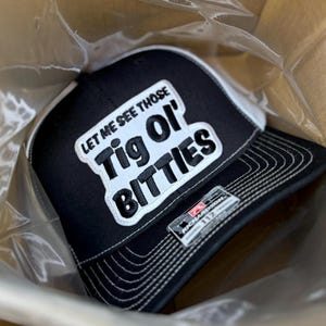Può includere: Cappellino da camionista nero e bianco con la scritta "LET ME SEE THOSE Tig Ol' BITTIES" in bianco. Il cappellino ha una visiera nera e un pannello frontale bianco. È un Richardson 112.