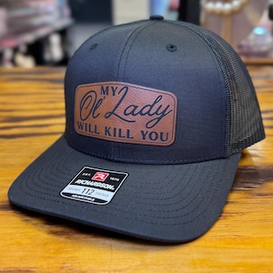 Könnte beinhalten: Schwarze Trucker-Cap mit Mesh-Rückseite und gebogenem Schirm. Auf dem braunen Lederpatch steht "My Ol' Lady Will Kill You". Die Kappe ist verstellbar.