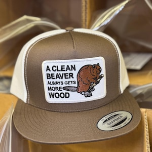 May include: A brown and white trucker hat with a patch that says "A Clean Beaver Always Gets More Wood" and a cartoon beaver holding a log. The hat has a black "The Authentic Snapback" label.