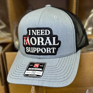 May include: Grey trucker hat with a black mesh back and a curved peak. The hat features a black patch with the words "I NEED MORAL SUPPORT" in white, with the word "MORAL" crossed out in red. A Richardson tag is visible.