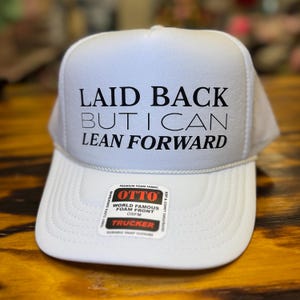 Puede incluir: Gorra de camionero blanca con el texto negro "LAID BACK BUT I CAN LEAN FORWARD". La gorra tiene una parte delantera de espuma blanca y una parte trasera de malla. Una pequeña pegatina en la visera dice "OTTO WORLD FAMOUS FOAM FRONT".