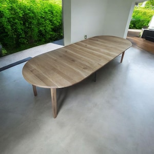 Può includere: Un grande tavolo da pranzo ovale in legno con finitura marrone chiaro. Il tavolo ha una superficie liscia ed è sostenuto da quattro gambe chiare. Le dimensioni del tavolo sono di circa 300 cm di lunghezza e 120 cm di larghezza.