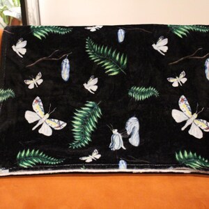 May include: A black throw blanket with a white, green, and blue butterfly and fern pattern. The blanket is folded over an orange couch. The butterflies have detailed wing patterns.