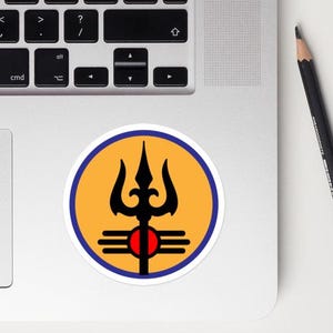 Puede incluir: Una pegatina redonda con un símbolo de tridente negro, un círculo rojo y líneas horizontales sobre un fondo amarillo, enmarcado por un borde azul. La pegatina está colocada en un ordenador portátil, sobre una superficie blanca.