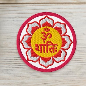 Könnte beinhalten: Gestickter Aufnäher mit einem rot-weißen Blumenmuster, mit einem gelben Zentrum mit dem Om-Symbol und dem Wort "Shanti" in Orange. Zwei kleine Sukkulenten in Töpfen sind ebenfalls zu sehen.
