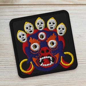 May include: Embroidered patch featuring a stylized, colorful demon face with multiple eyes and skulls. The patch is square with a black background, red, blue, and yellow accents. Two small potted succulents are in the background.