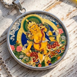 May include: A silver-toned metal pendant necklace featuring a circular frame containing a colorful illustration of a yellow deity figure against a blue and green background with floral accents, suspended from a matching silver-toned box chain.