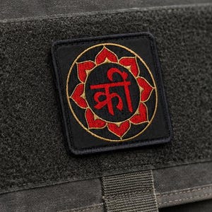 May include: A dark olive green tactical messenger bag featuring a rectangular flap with a black zipper pocket and a central black patch. The patch displays a red lotus flower design with the Devanagari script character 'क्रीं' in the center.