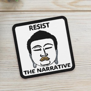 May include: A square patch with the text "RESIST THE NARRATIVE" and a graphic of a serene face with closed eyes and earrings. Two small potted succulents are also in the image, set on a light wood surface.