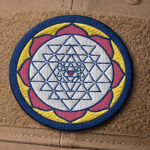 May include: A tan baseball cap with a circular patch featuring a geometric design. The patch has a blue border, yellow accents, and a central pattern of white triangles and a red dot.