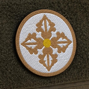 Puede incluir: Un parche bordado circular con un diseño geométrico simétrico en marrón y blanco con un punto amarillo central, fijado a una bolsa táctica de color verde oliva oscuro con un panel rectangular de velcro para sujetar equipo adicional.
